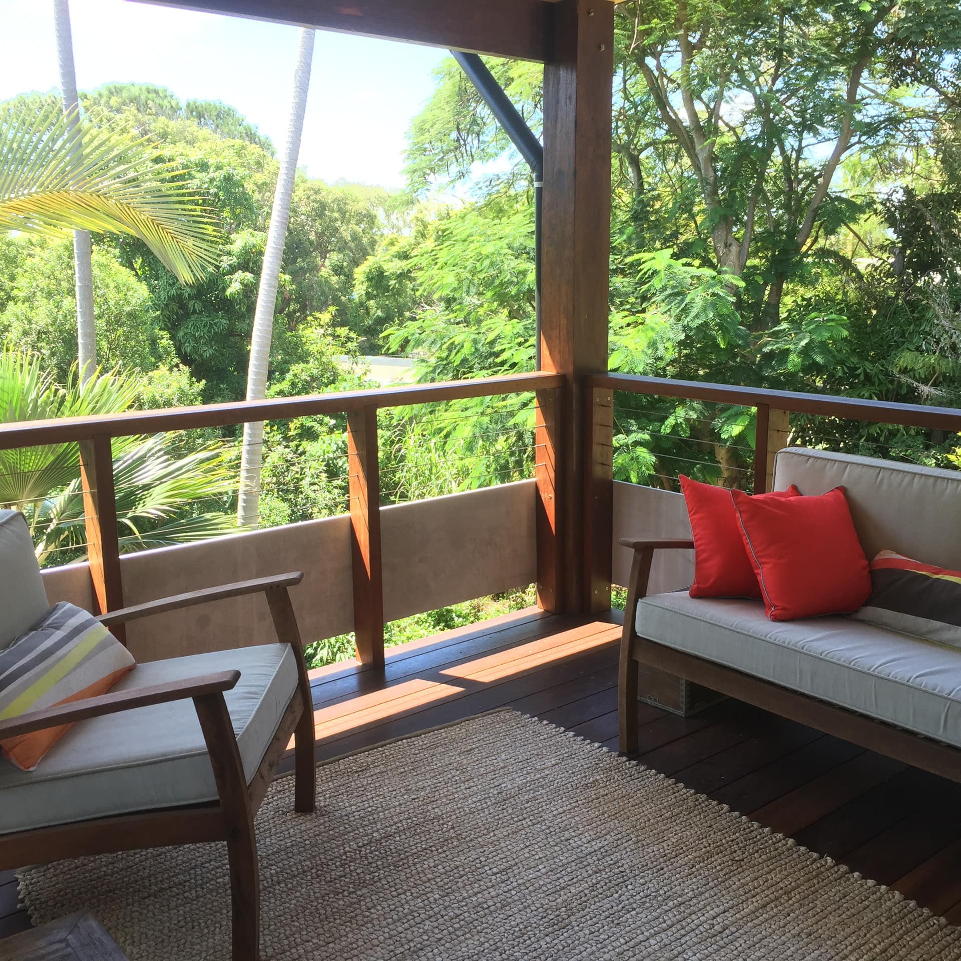 Wooden deck with tan canvas sofa and chair, colorful throw pillows, woven area rug, surrounded by lush greenery.