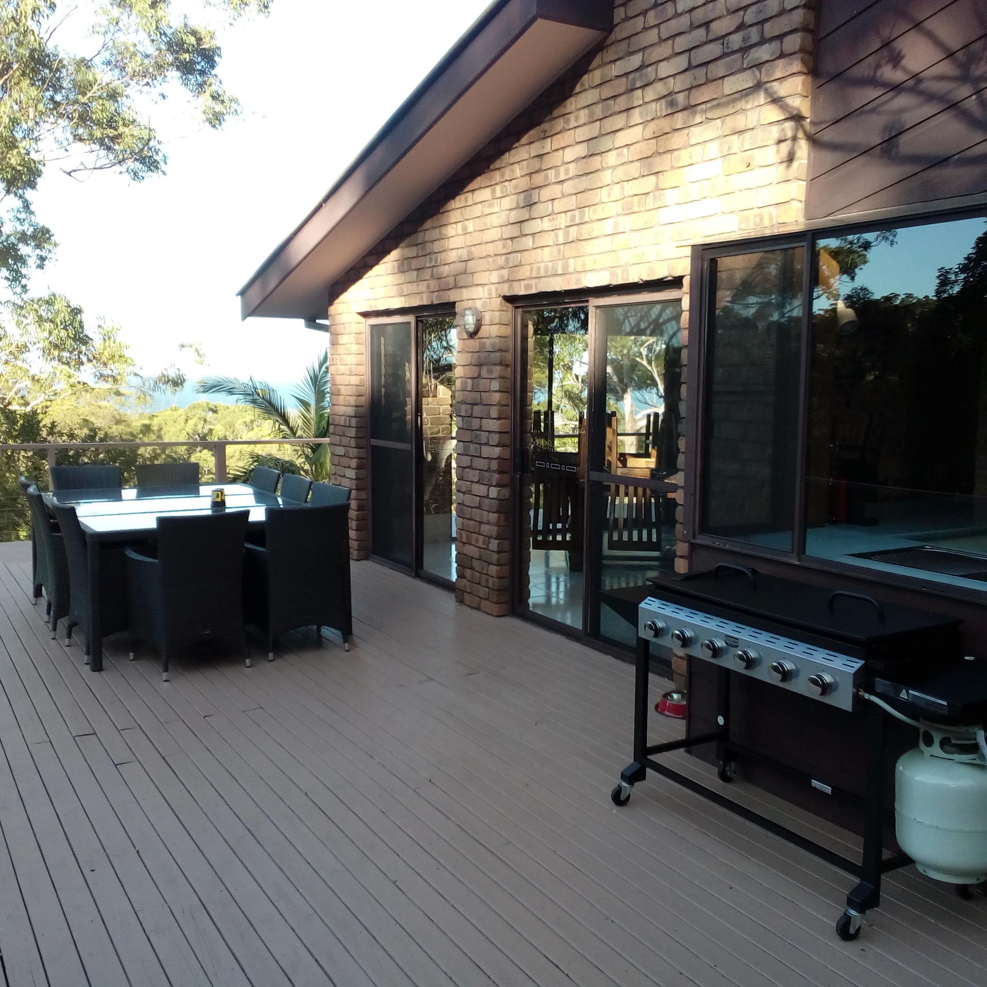 A large tan deck with views of surrounding vegetation, a black table with seating for ten, and grill.