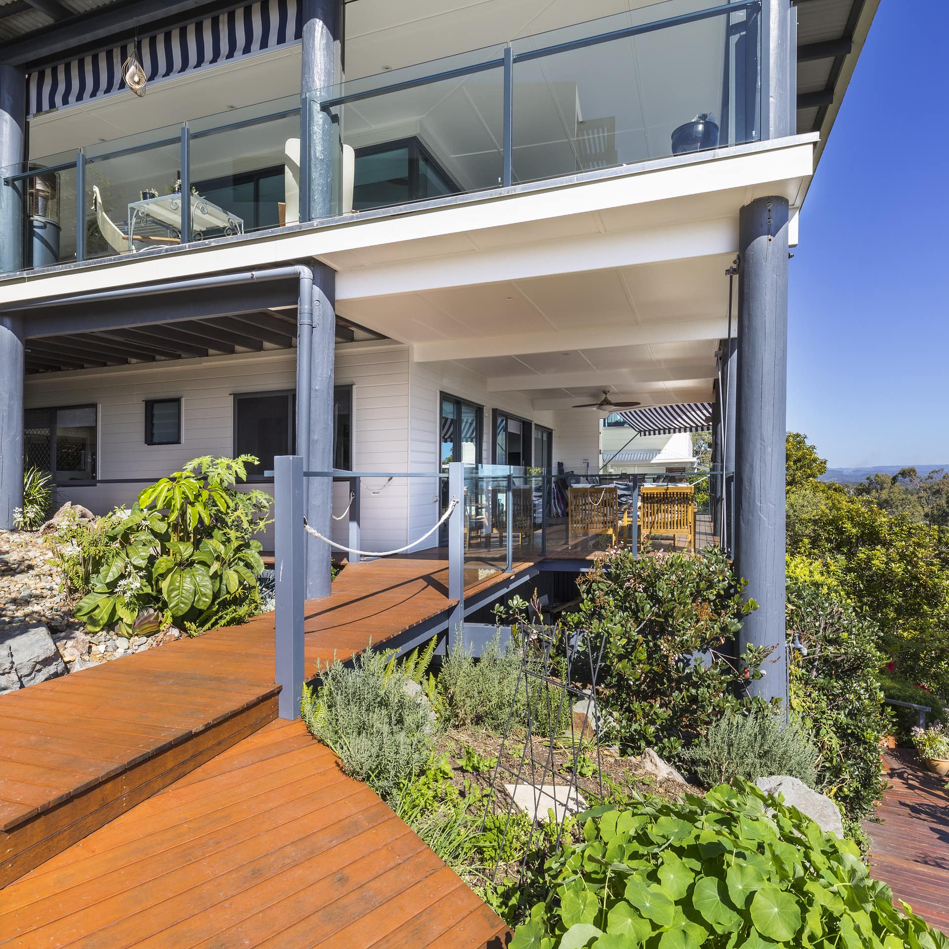 Exterior view of modern two-story home, raised wooden walkway, covered decks on both levels, surrounded by lush lawn.