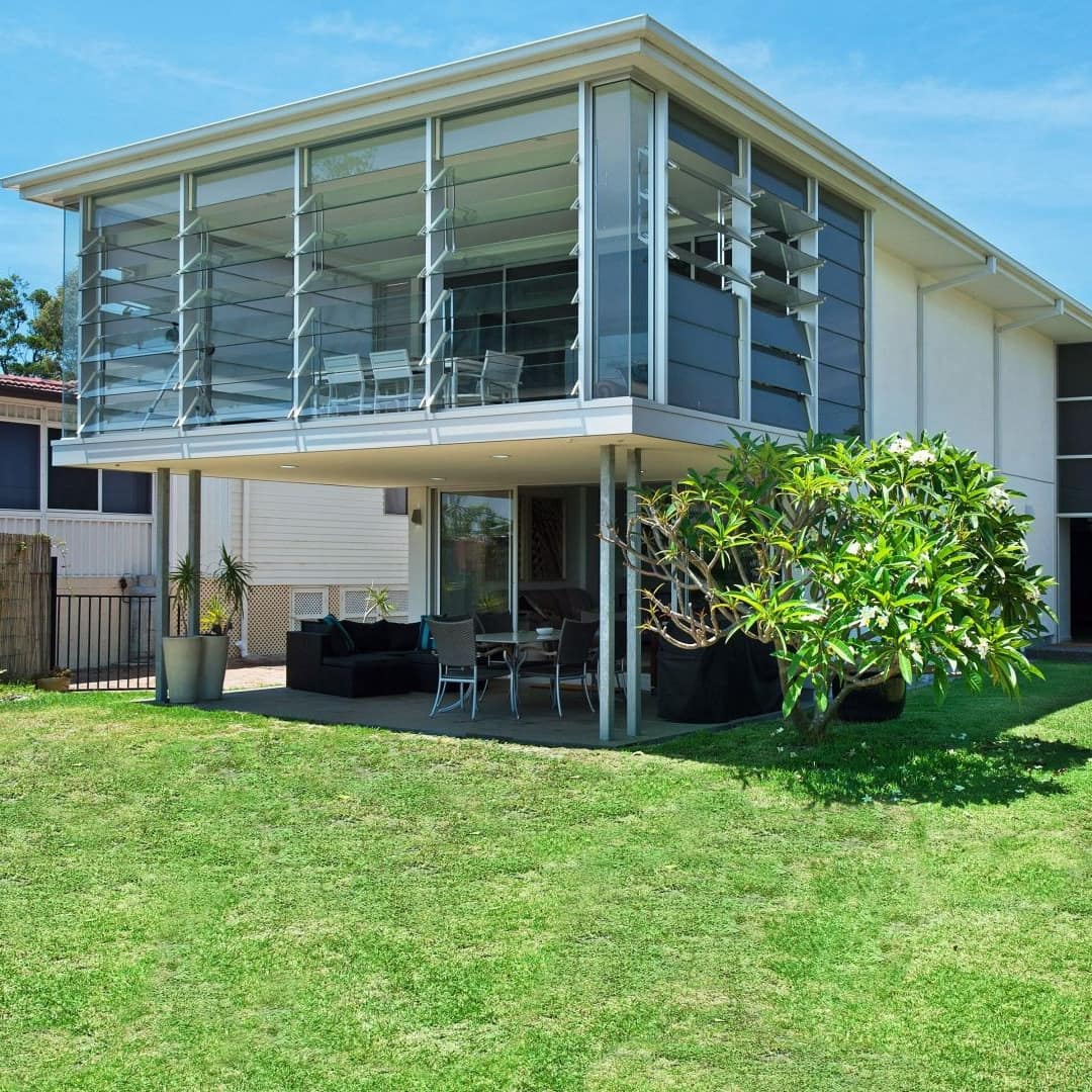 Modern beach house with lush lawn, floor to ceiling vented windows in the second story sunroom, and a patio below.