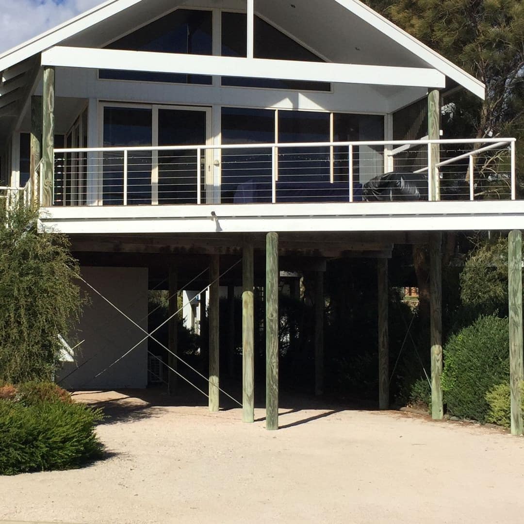 A raised beach house with a wrap-around verandah, white railings, and lush greenery surrounding the home.
