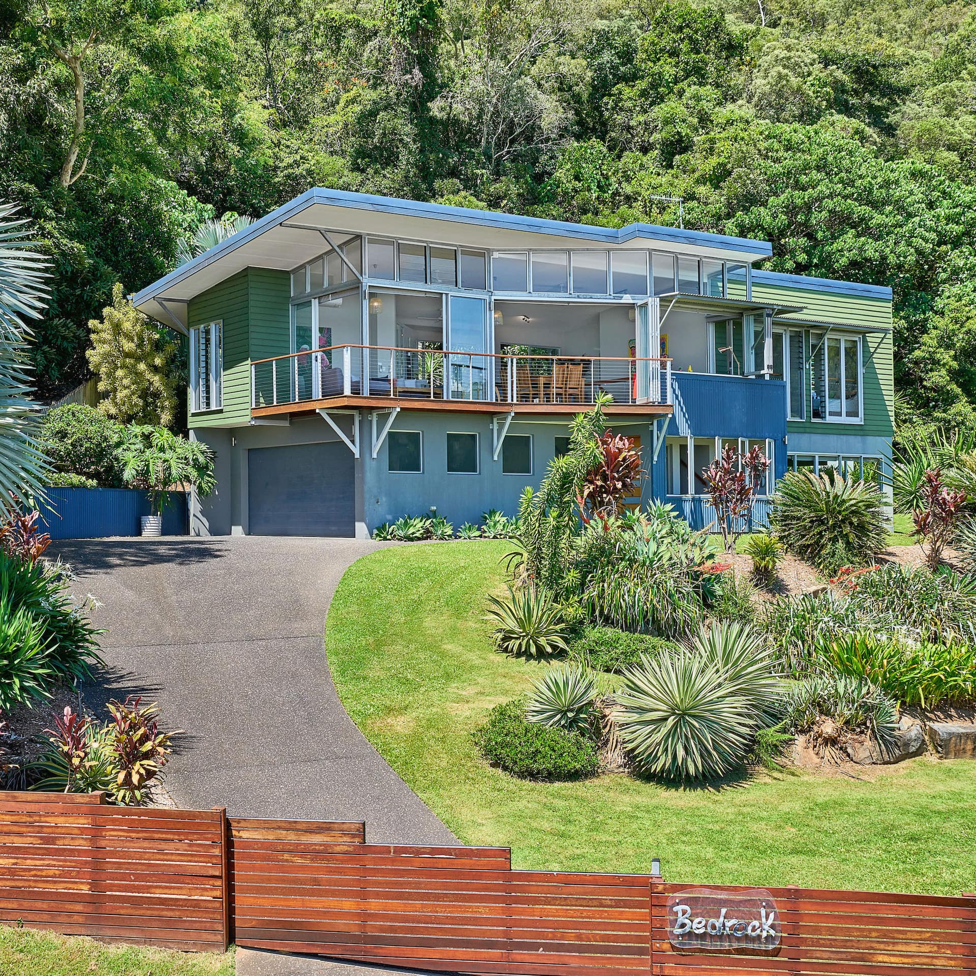 Exterior view of modern, beach home with green vinyl siding, balcony, expansive windows, and lush, landscaped lawn.
