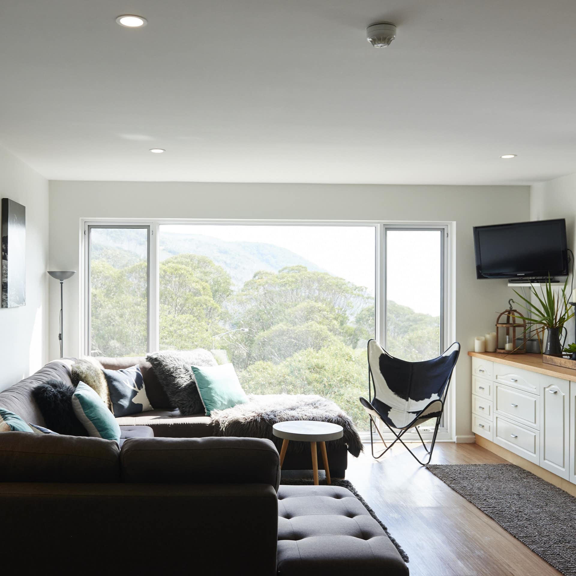 Sunlit living room with hardwood floors, grey sectional, flatscreen TV, butterfly chair, wide windows with mountain views.
