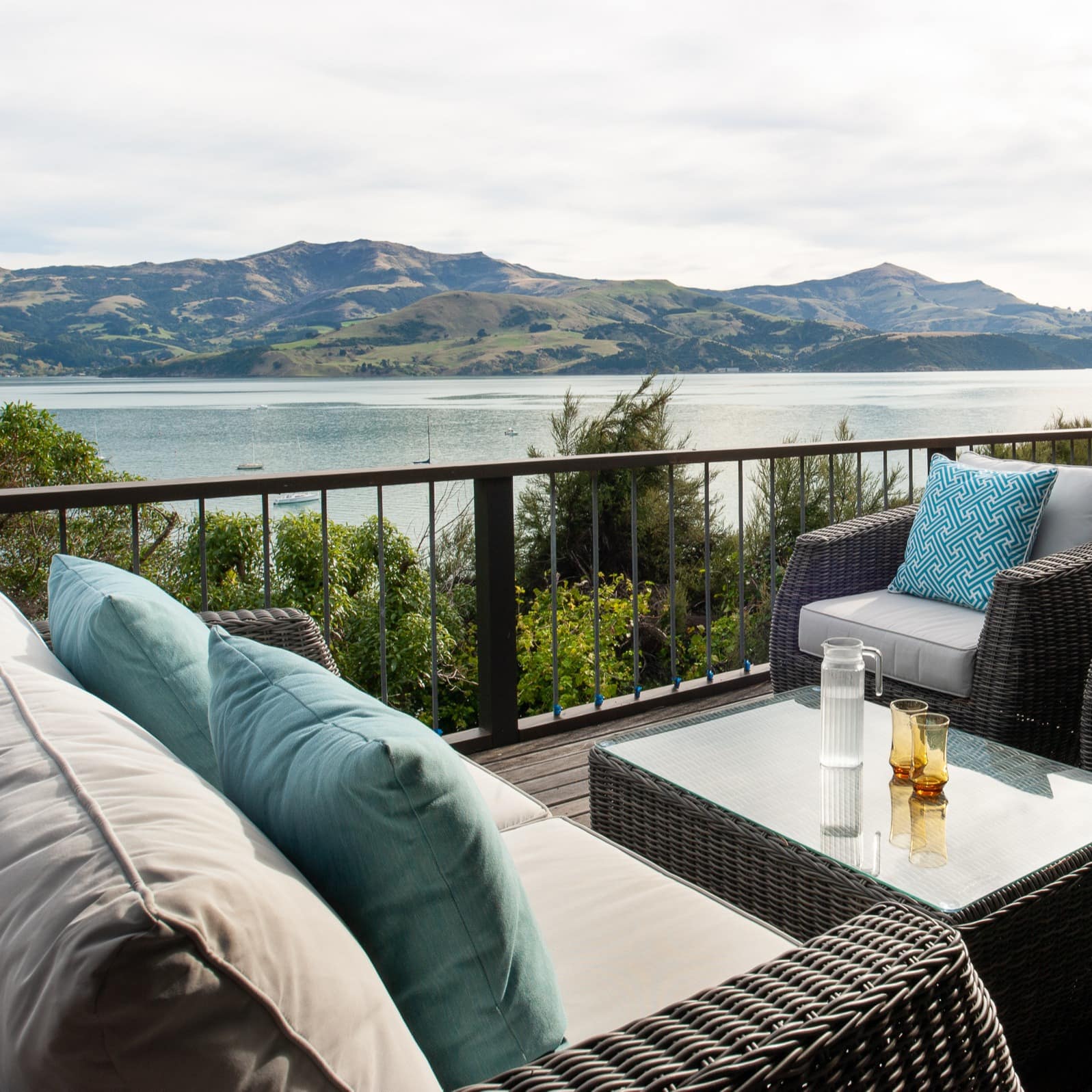 A wooden deck with black railings, matching grey wicker furniture, overlooking the harbour and mountain range.