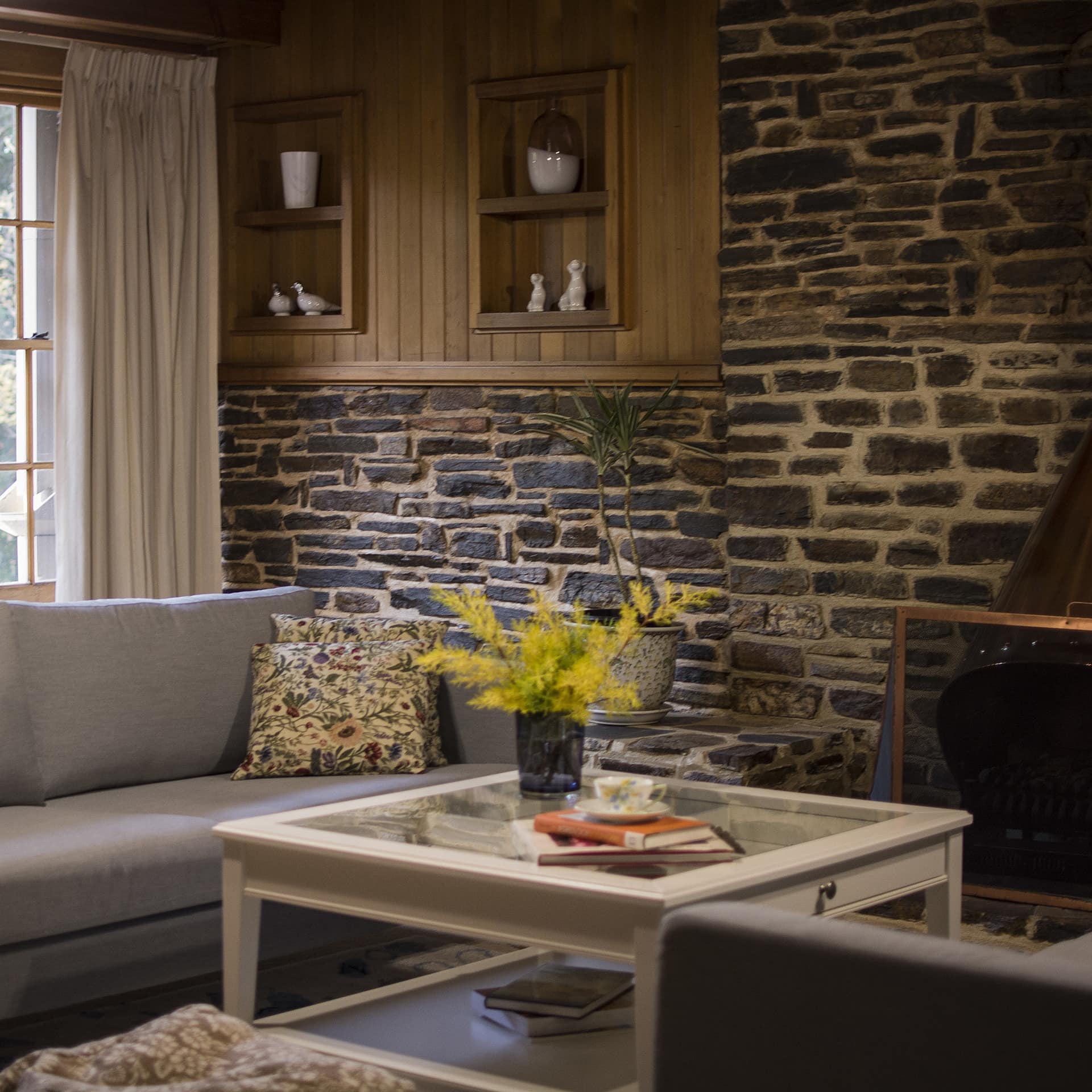 Living room with stone walls, inset wood shelves, grey canvas sofa, glass coffee table, wide windows with nature views.