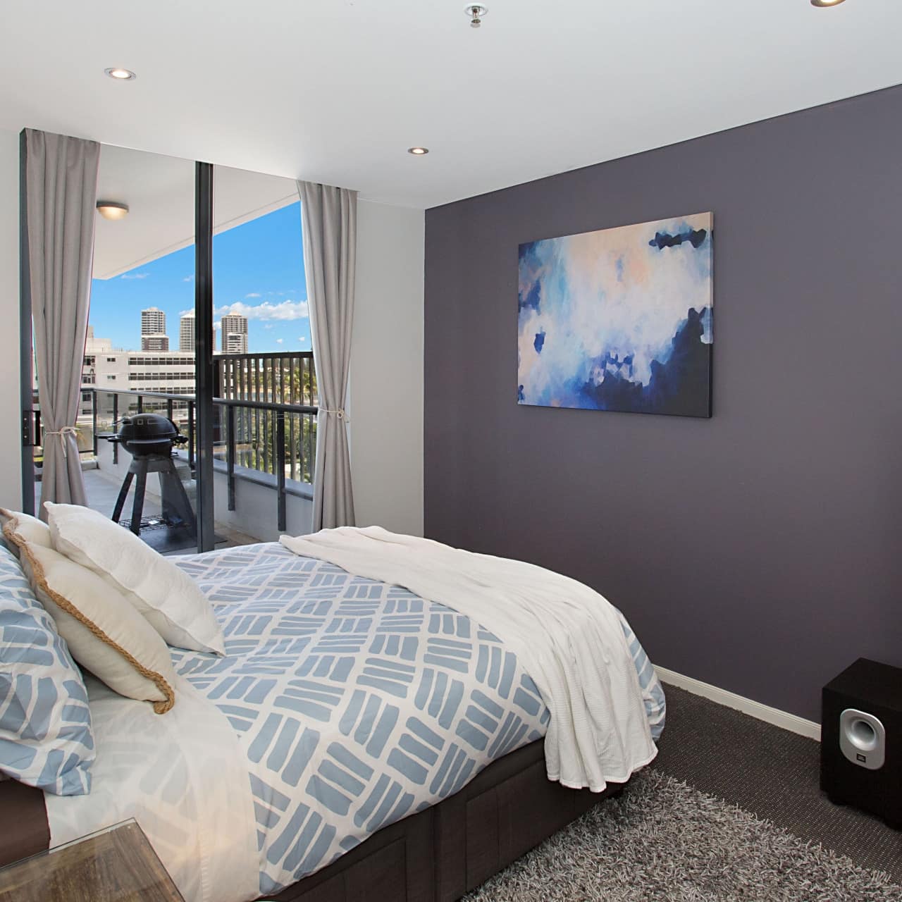 Grey bedroom with queen-sized bed, fine, blue-patterned linens, wood chest and sliding doors to balcony with grill