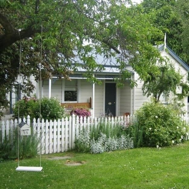 A white cottage in Arrowtown with flower bushes growing over a picket fence and a tree swing in the foreground