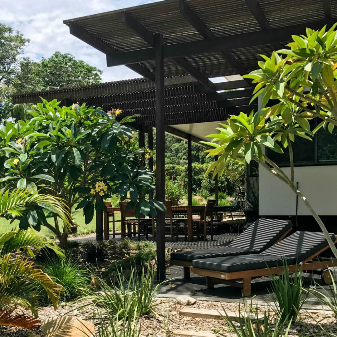 A covered patio space with black lounge chairs, surrounded by a lush tropical garden.