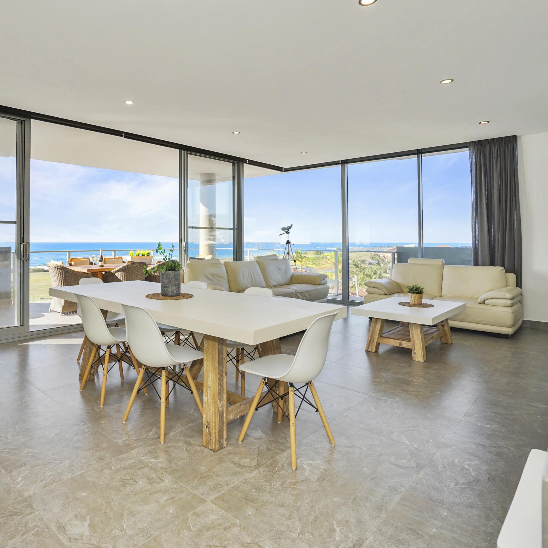 Open living and dining room, floor to ceiling windows with ocean views, beige tile floor, modern beige furnishings.