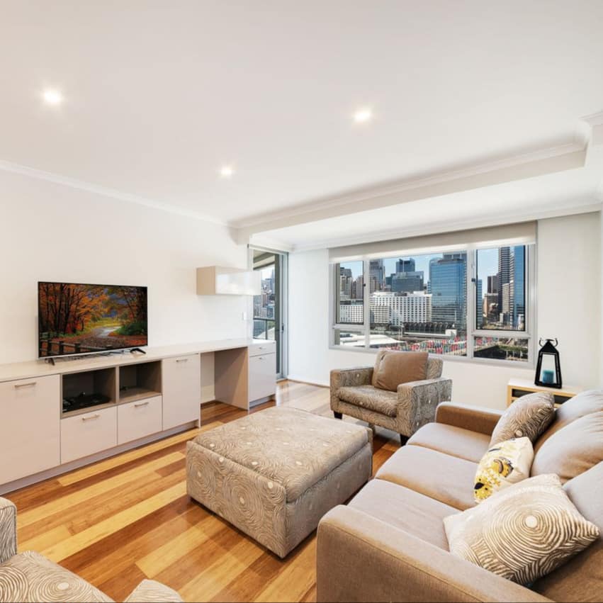 Open living room with hardwood floors, white walls, beige sofa and chairs, flatscreen TV, and wide windows.
