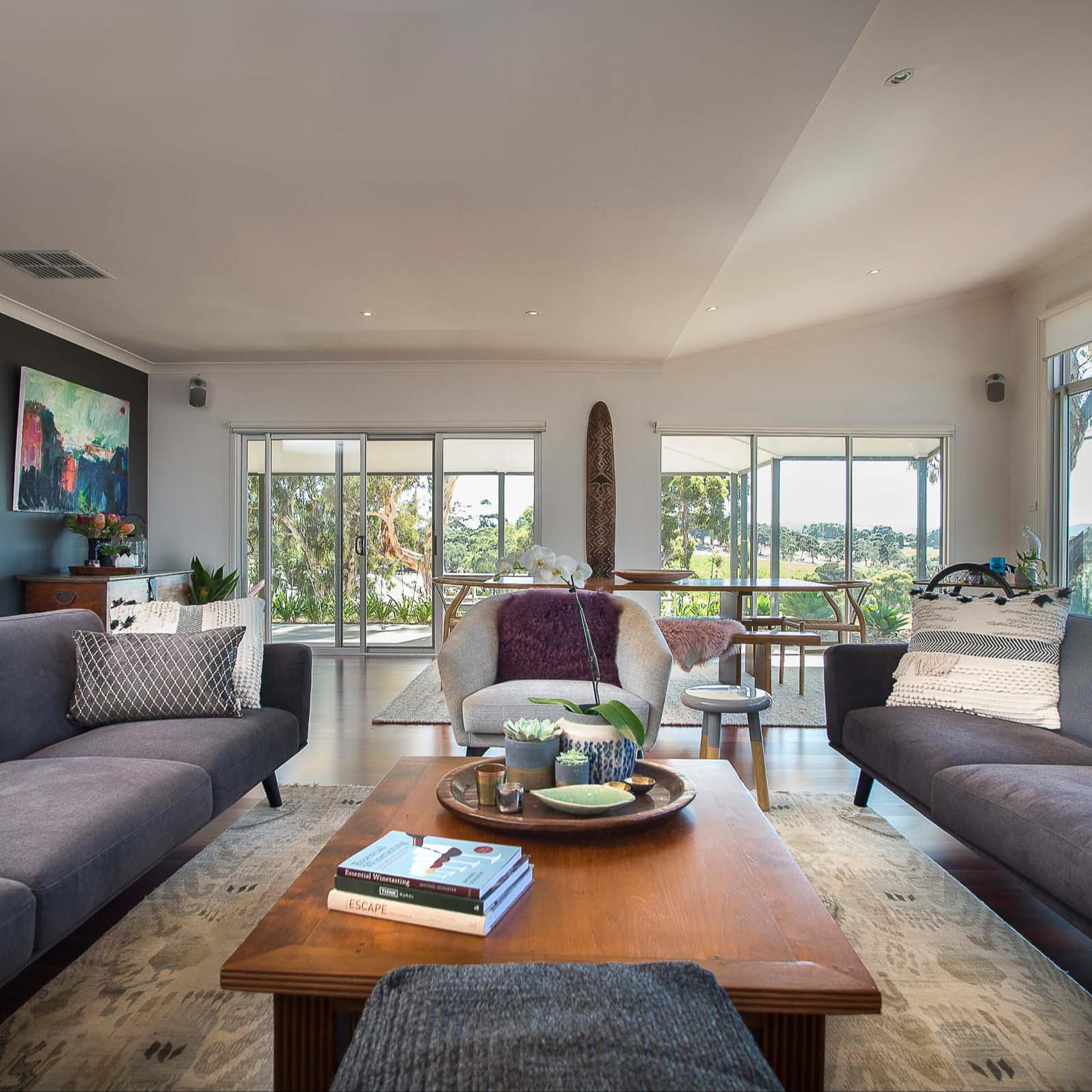 Modern living and dining room with grey sofas, white chair, wooden coffee table, wide windows with panoramic vineyard views.