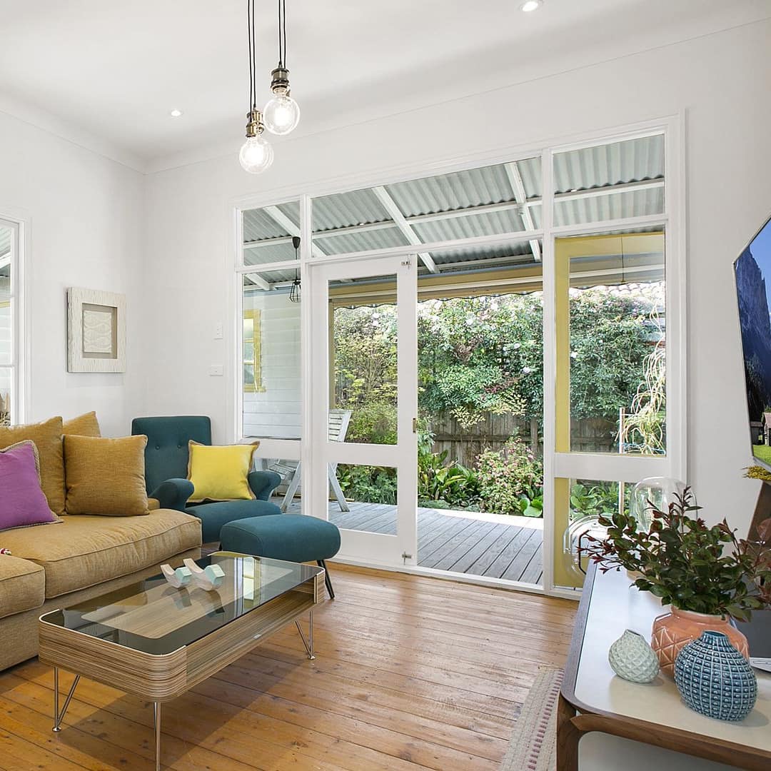 Living room with hardwood floors, tan sofa, green and teal chairs, glass coffee table, flatscreen TV, tall windows.