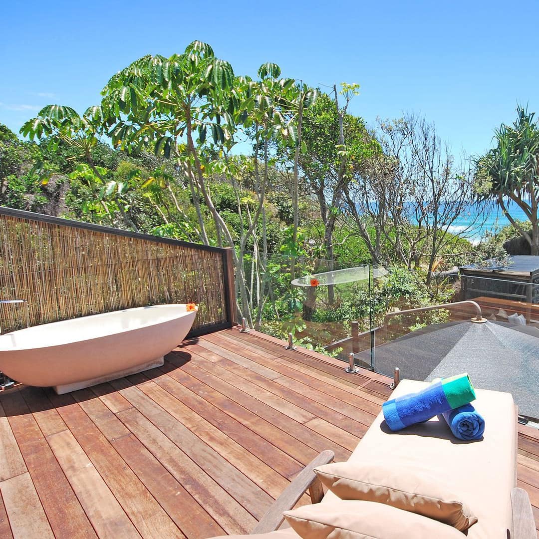 A wooden deck with tan lounge chair and outdoor tub, overlooking lush vegetation and the beach.