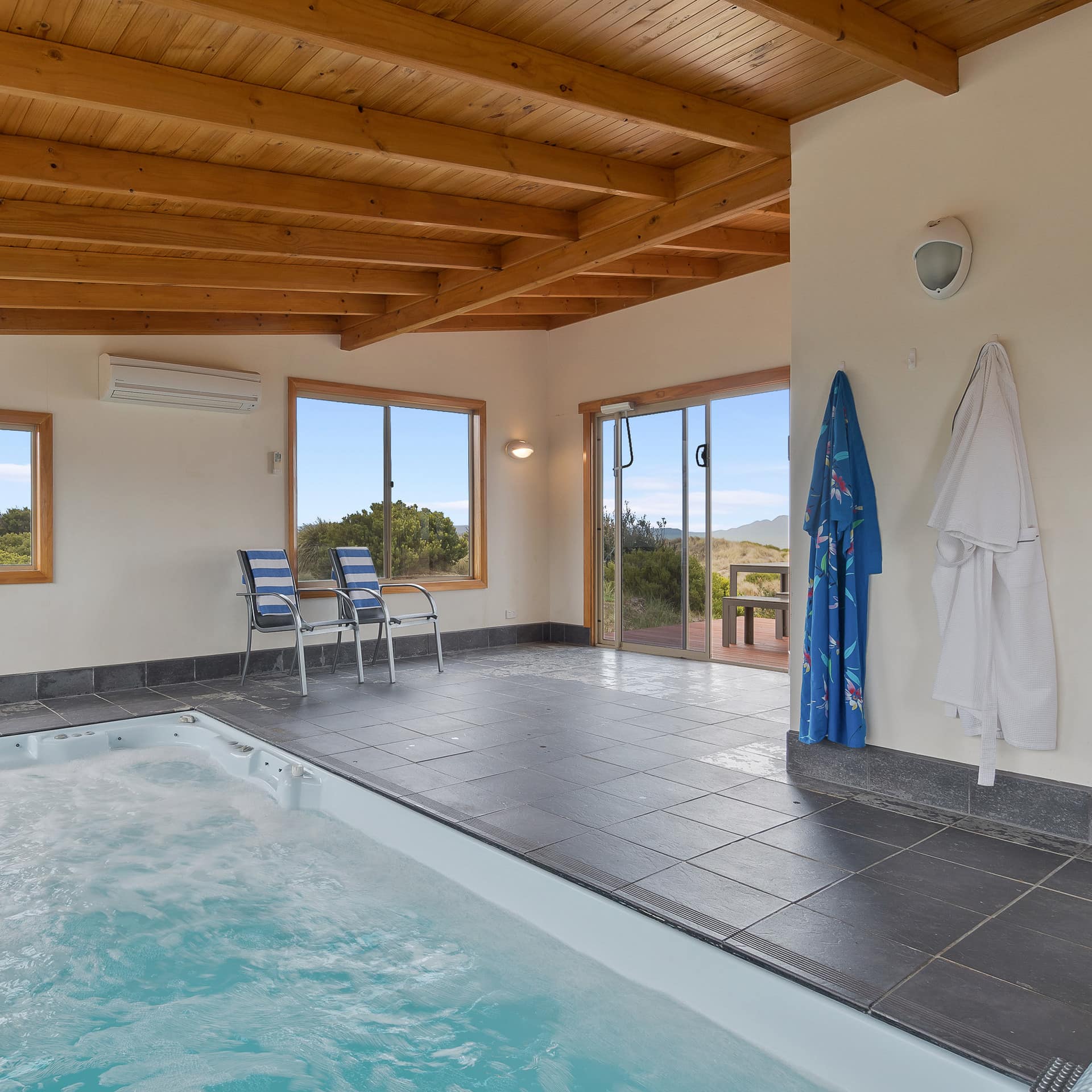 View of a private indoor pool with Jacuzzi jets, grey tile floors, vaulted ceiling, and robes hanging on the wall.