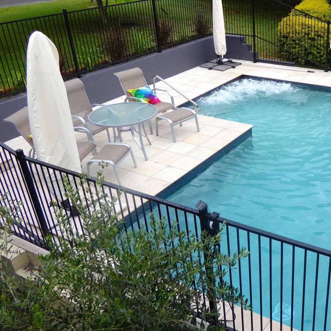 A square inground pool with beige tile patio space, tan lounge chairs, enclosed by black iron fence and manicured lawn.
