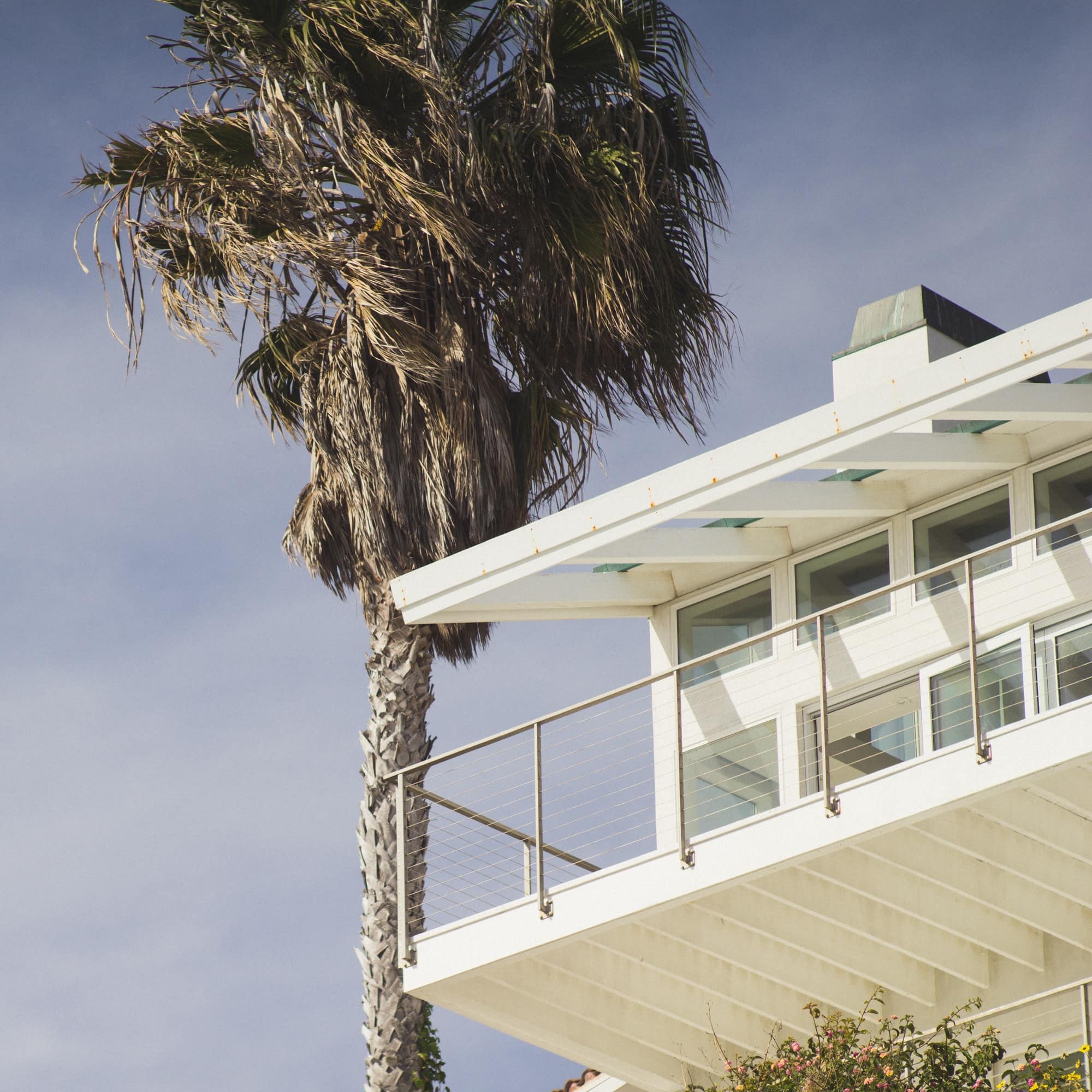 White, modern luxury beach house with upper level balcony, many windows and a palm tree set against a soft blue sky