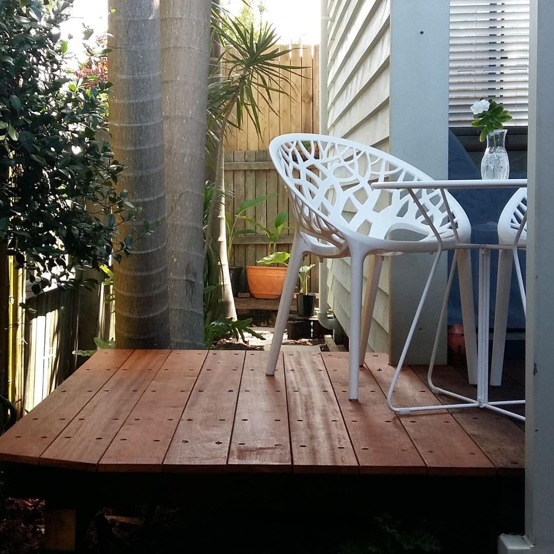 View of a wooden deck with a white bistro dining set, surrounded by bamboo and palm trees.