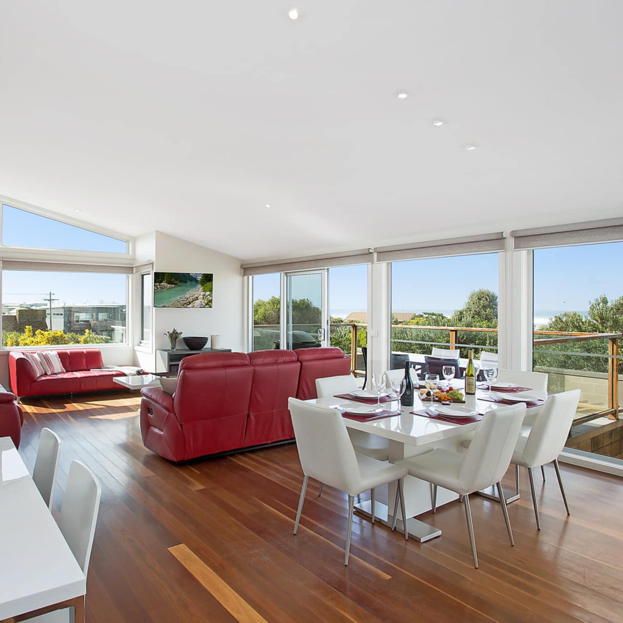 Open living and dining room with hardwood floors, floor to ceiling windows, white dining set, and red leather sofas.