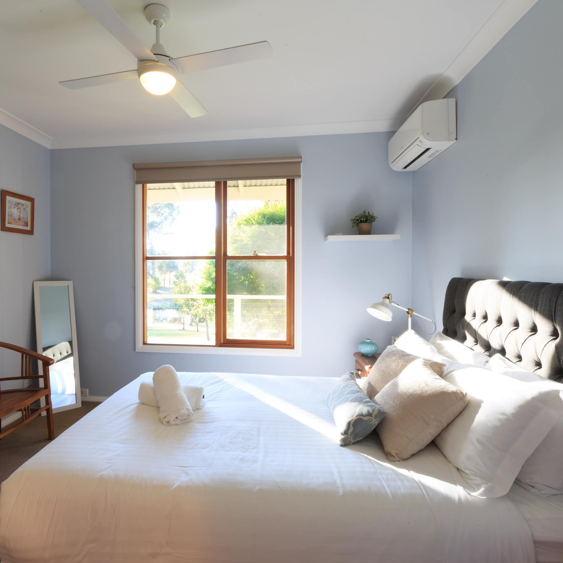 Sunlit bedroom with light blue walls, a king-size bed, black fabric headboard, white bedding, wooden bench.