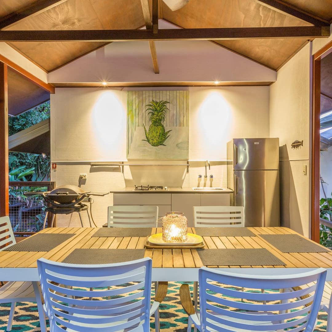 Outdoor kitchen on covered deck with wood dining set, wood floors, and tropical plants.