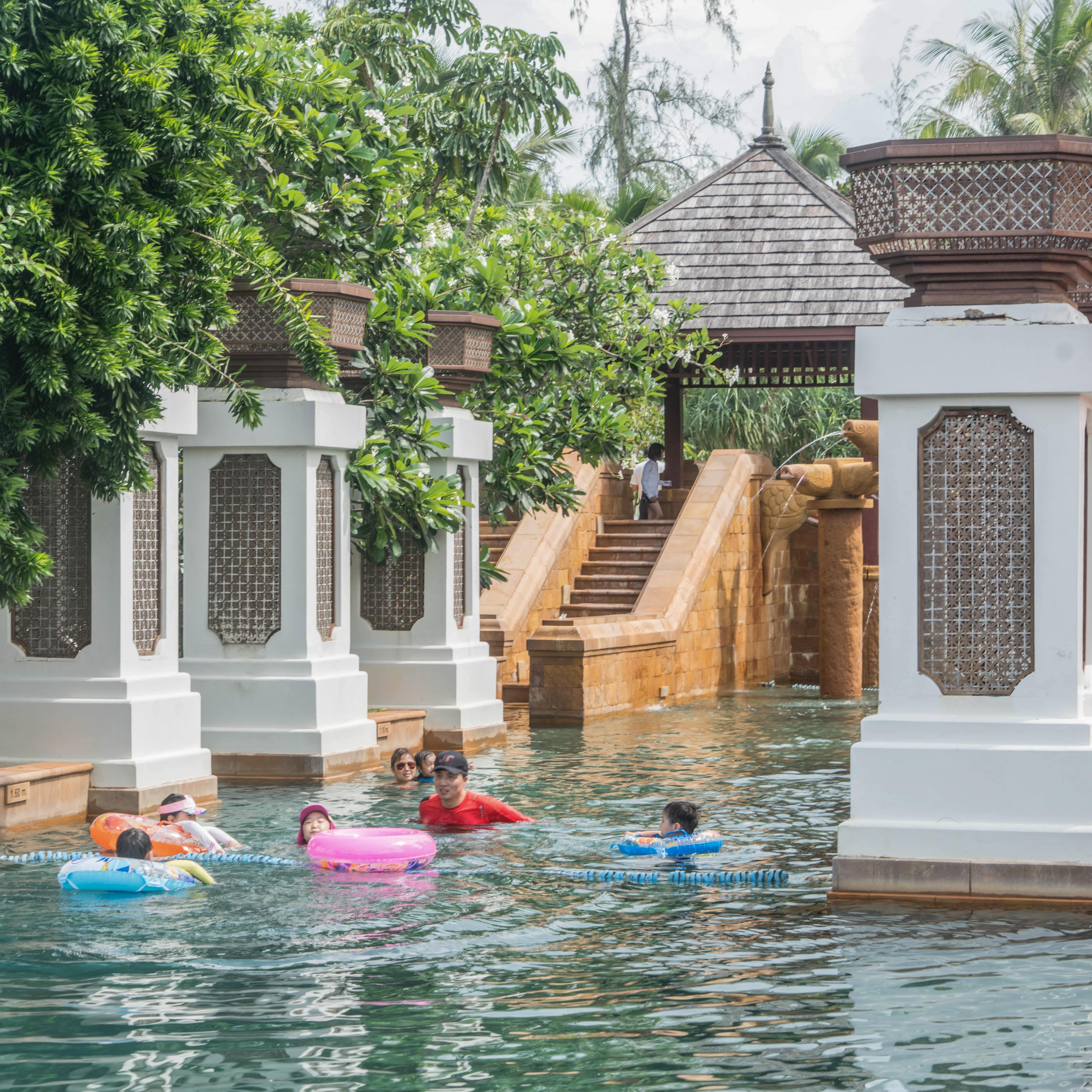 Children playing in exotic pool of Phuket villa with brown and white columns and stairs to gazebo in background