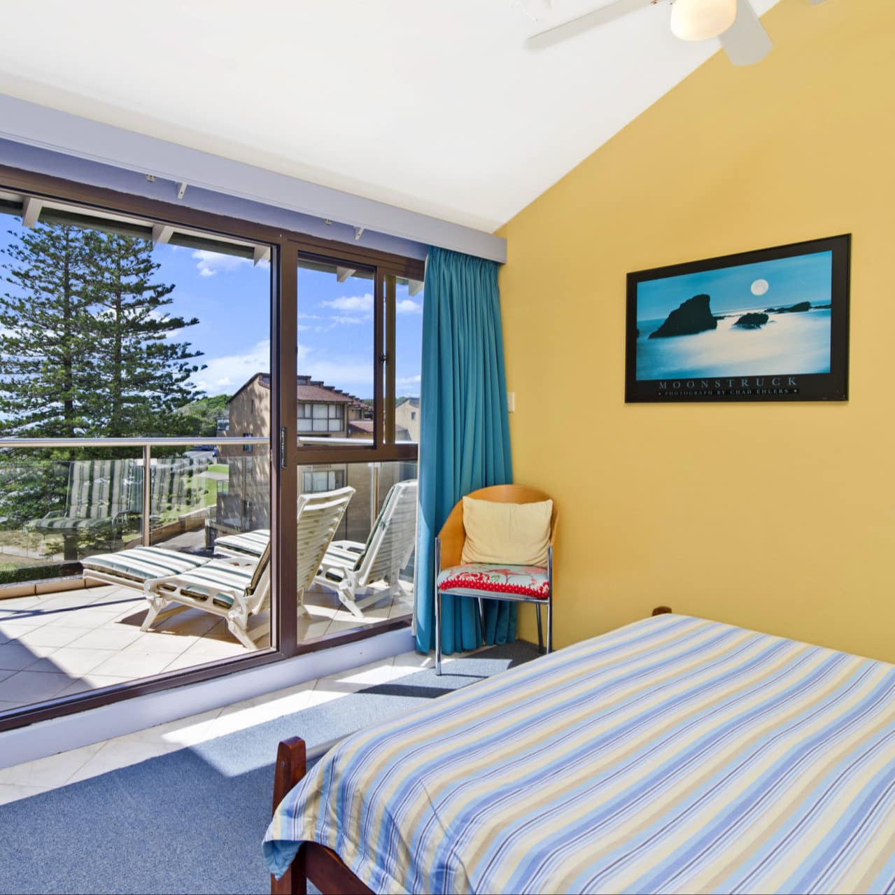 Brightly coloured room with bed facing floor-to-ceiling windows and sliding door to patio overlooking trees, sea and blue sky