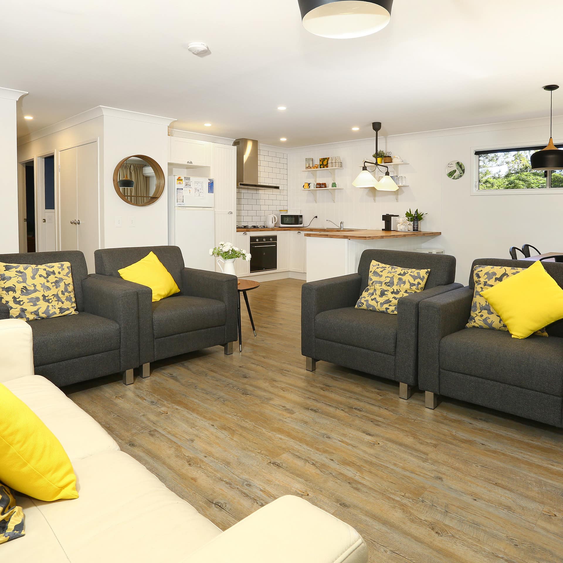 Open living room and kitchen with hardwood floors, grey chairs, tan sofa, yellow throw pillows, and modern kitchen.