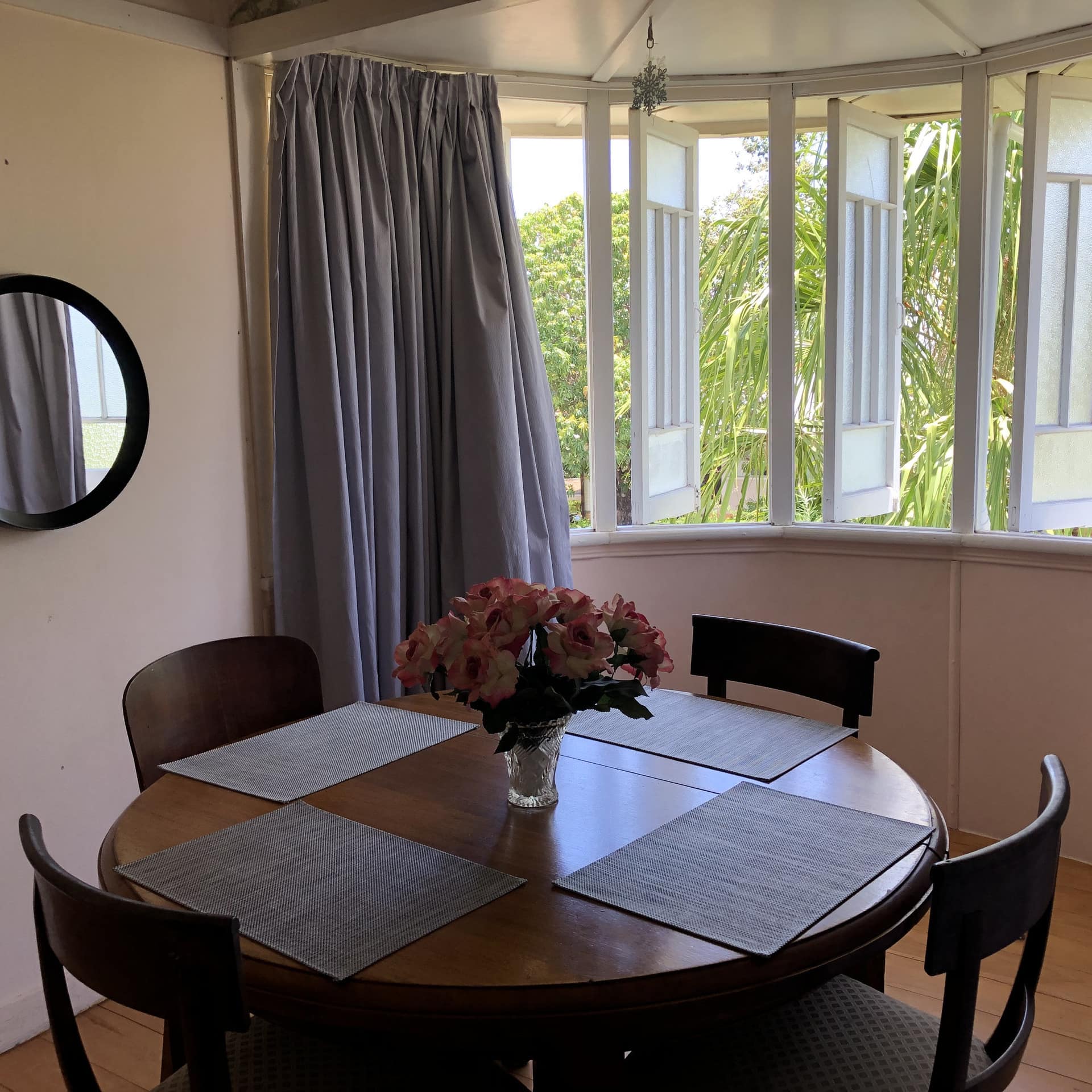 Dining room with round, wooden dining table with a vase of roses, open bay window with views of tropical greenery.