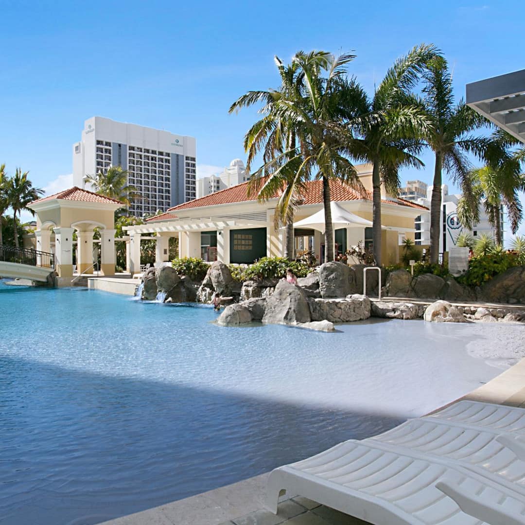 Sunny view of resort pool with beige tile pool deck, white lounge chair, cabanas, and palm trees.