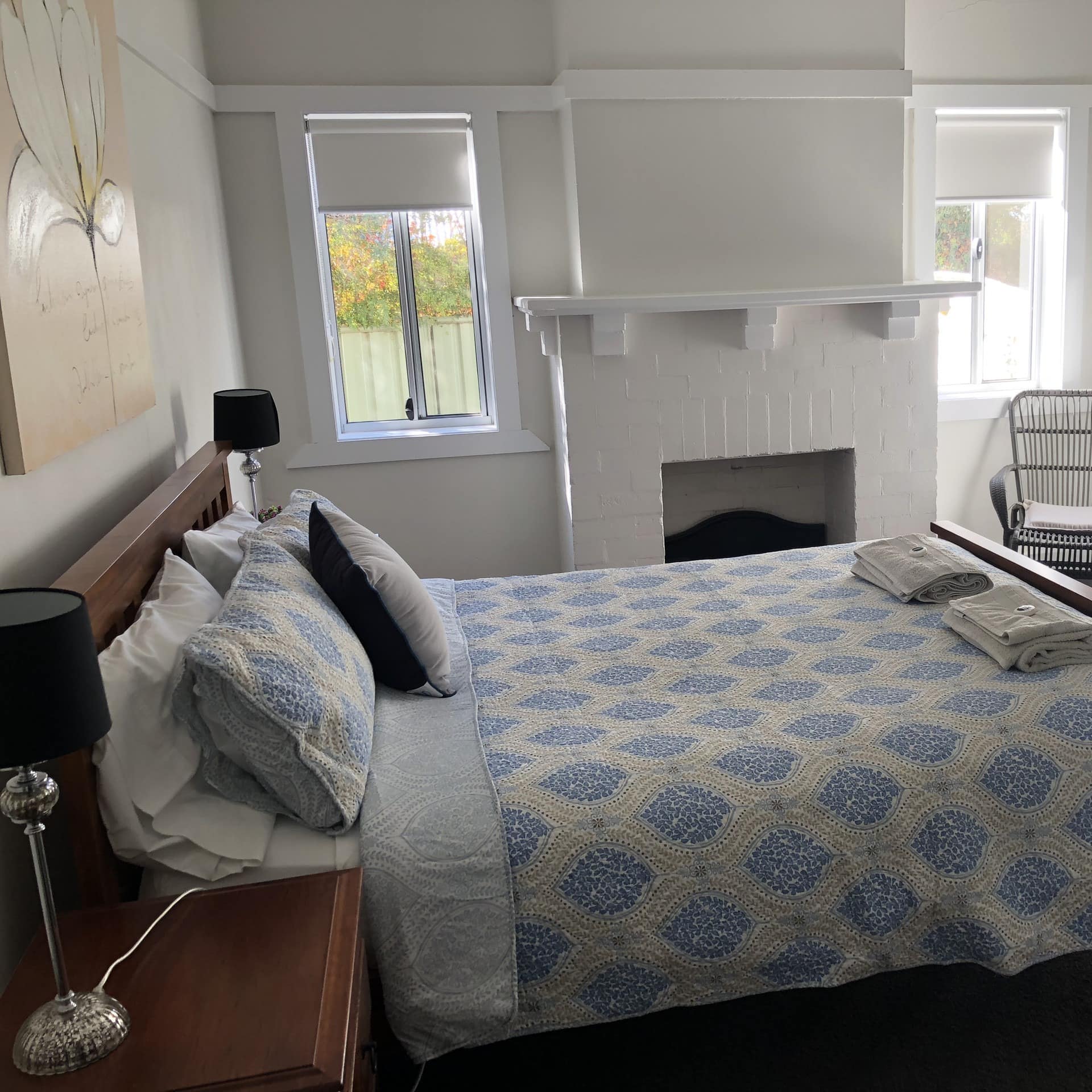King-size wood-frame bed and blue and white floral bedding in bright, white room with high ceiling and white brick fireplace