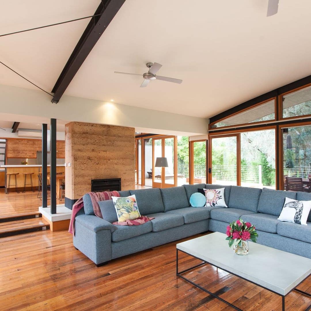 Spacious open kitchen and sunken living room, hardwood floors, fireplace, and L-shaped blue sofa, floor to ceiling windows.