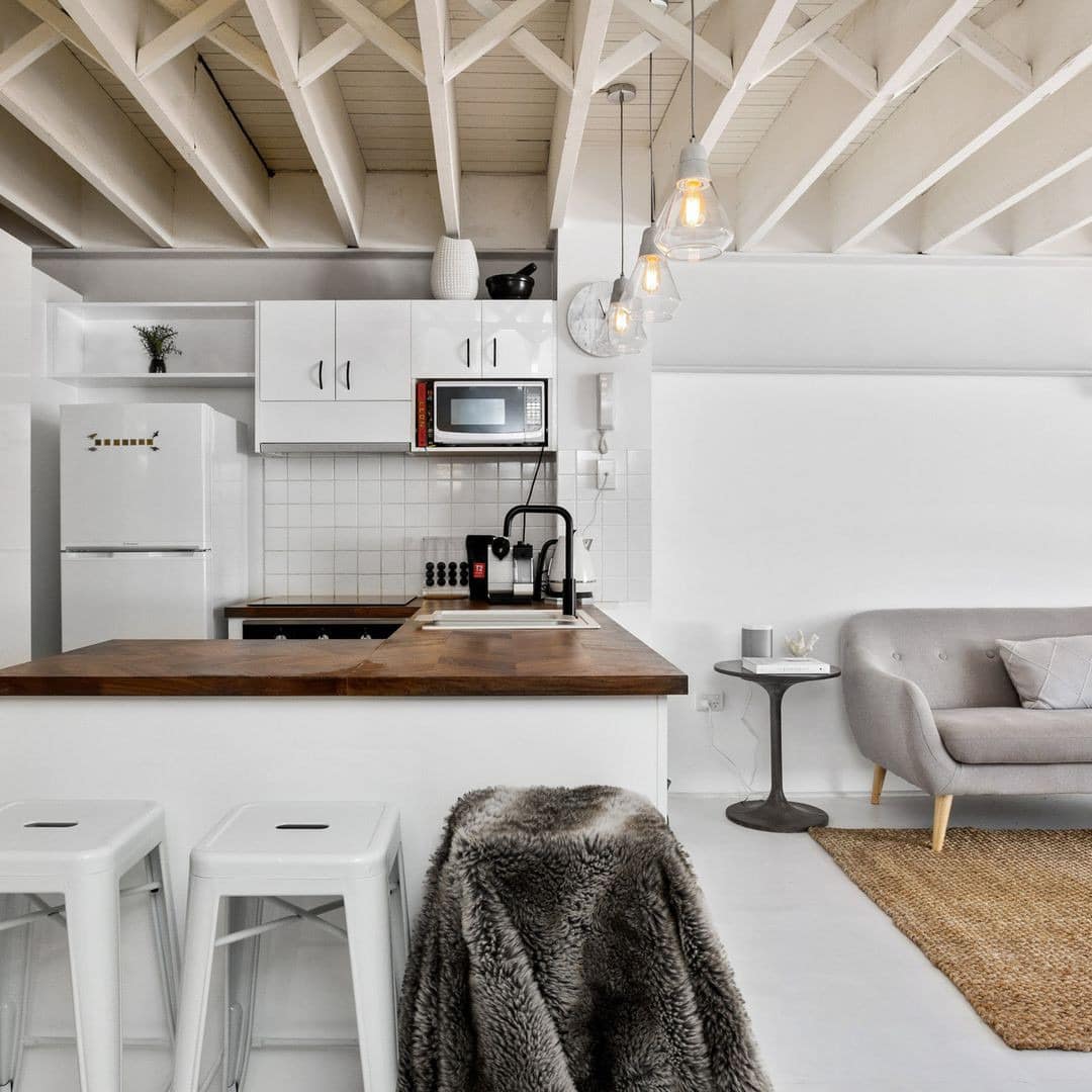 Minimalist open kitchen and living room with white cabinets, wooden countertops, grey sofa, and a tan woven rug.
