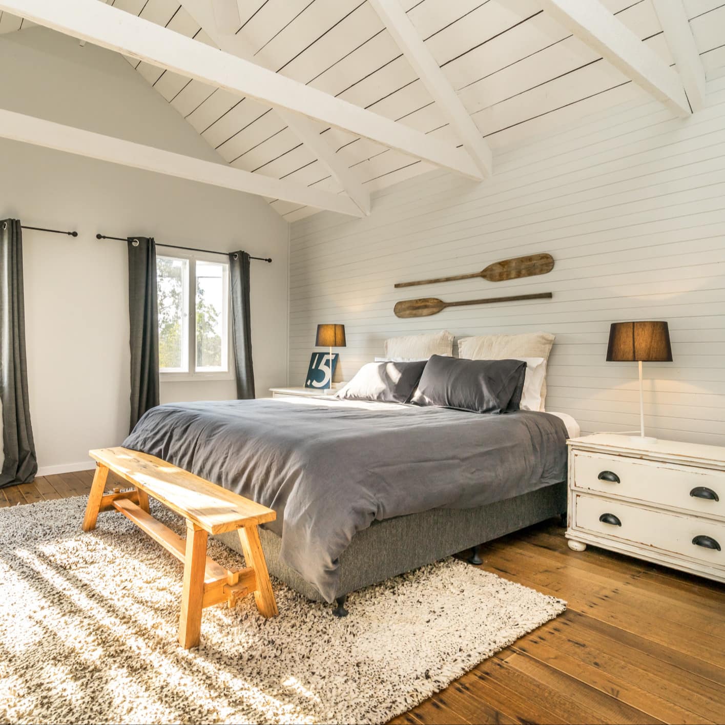 Sunlit bedroom with hardwood floors, vaulted ceiling with exposed beams, king size bed, grey bedding, shag area rug.