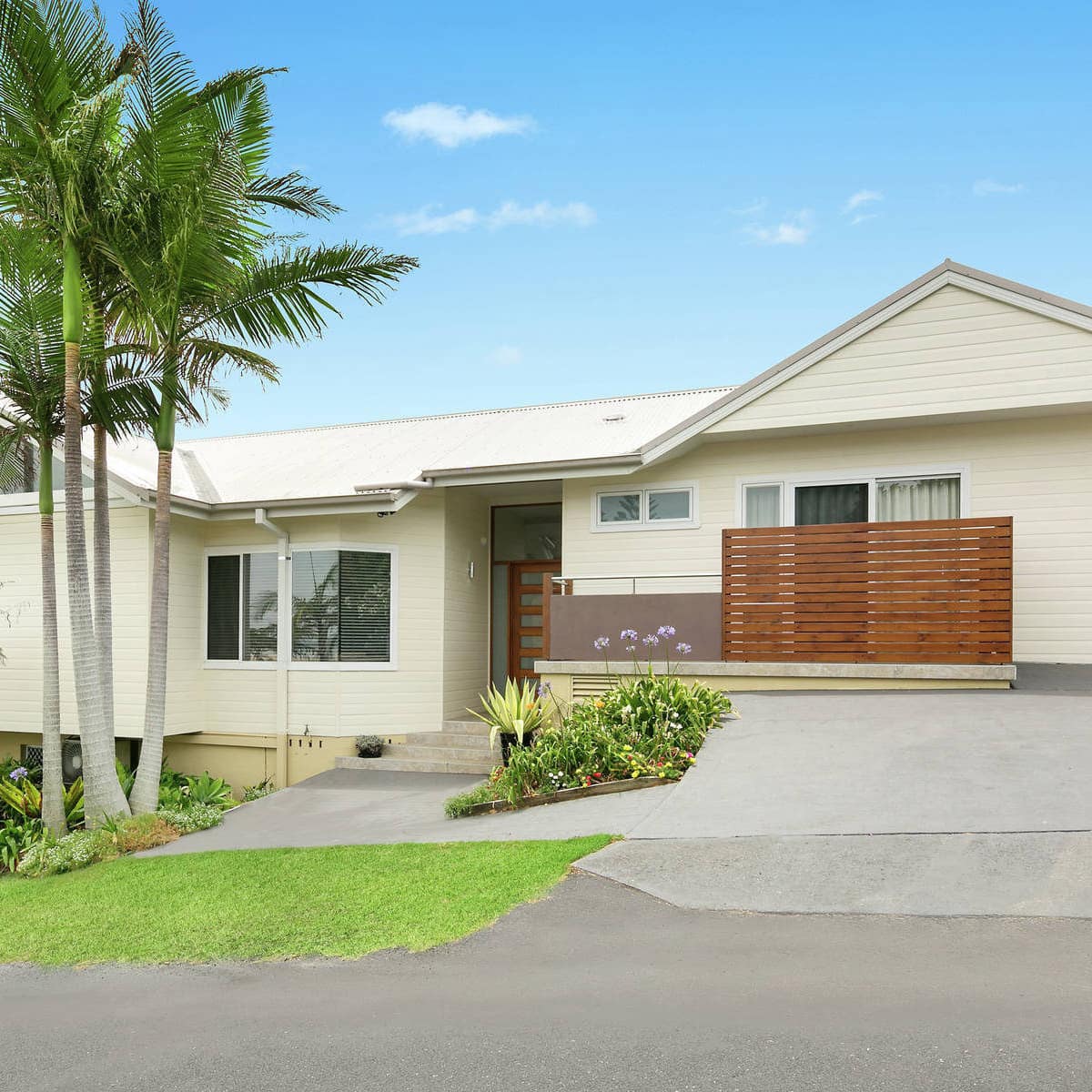 Exterior view of ranch style home, beige siding, wood door and railing, palm trees in the front yard, and a manicured lawn.