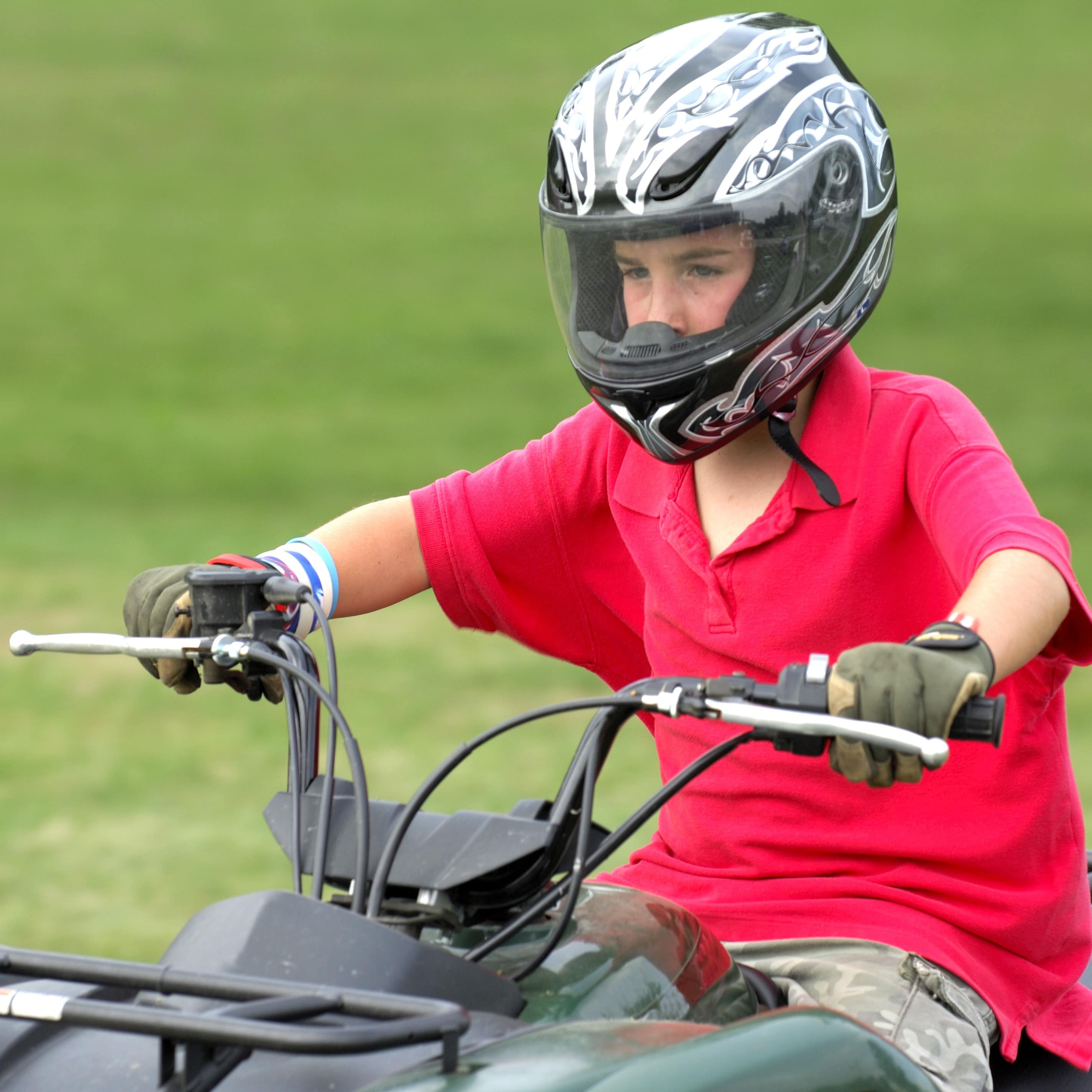 Boy with red shirt and black and white helmet riding on ATV with green grass in background
