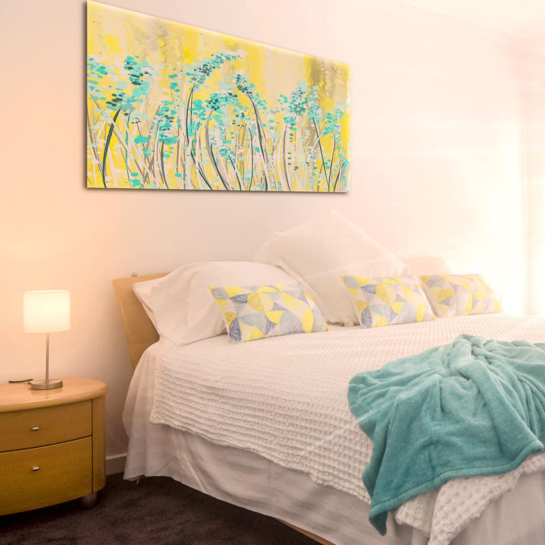 A sunlit room with a king size bed, white bedding, yellow and teal decorations, grey carpet, and white walls.