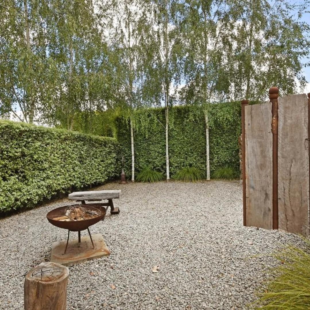 View of a rustic backyard space with pebble gravel, a firepit, tree stump seats secluded within walls of manicured shrubs.