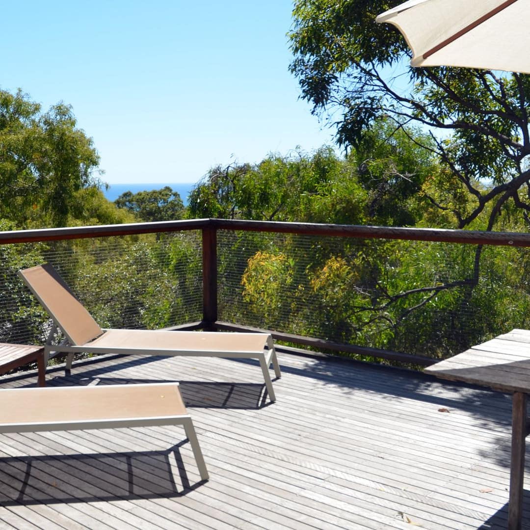 Sunny view of a wood veranda encircled by thick forest with two canvas lounge chairs and a wood dining set with umbrella.
