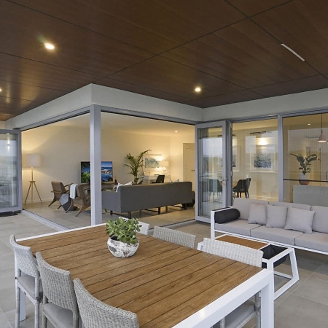 Spacious, covered veranda with grey tile, wood dining table, beige sofa and wicker chairs, and folding glass doors.
