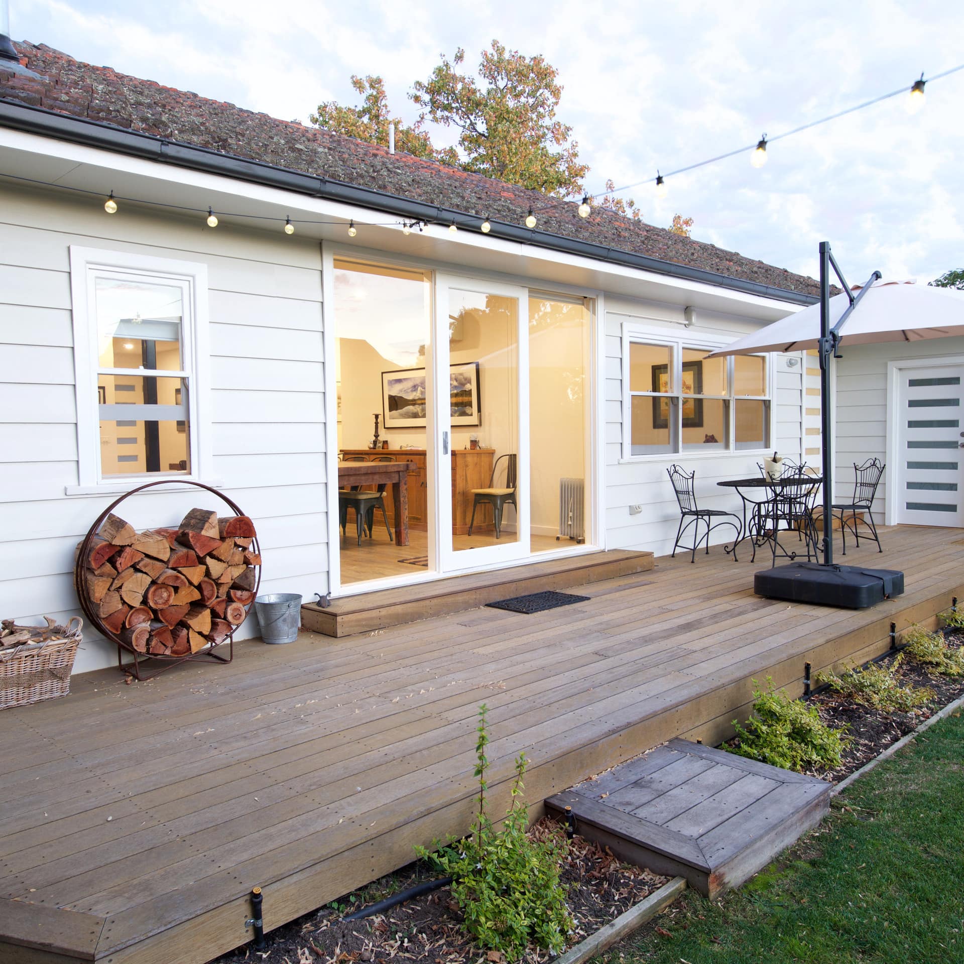 View of wooden deck of white cottage with black metal dining set, plant beds, stacked chopped wood and sliding glass doors.