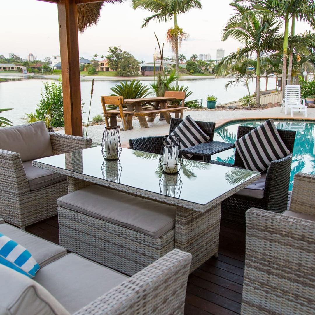 A patio space with wicker furniture next to a small inground pool, with views of a canal and surrounded by palm trees.