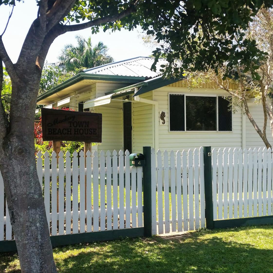 Exterior of 1-storey beach house with white siding, green metal roof, and white picket fence with overhanging tree.