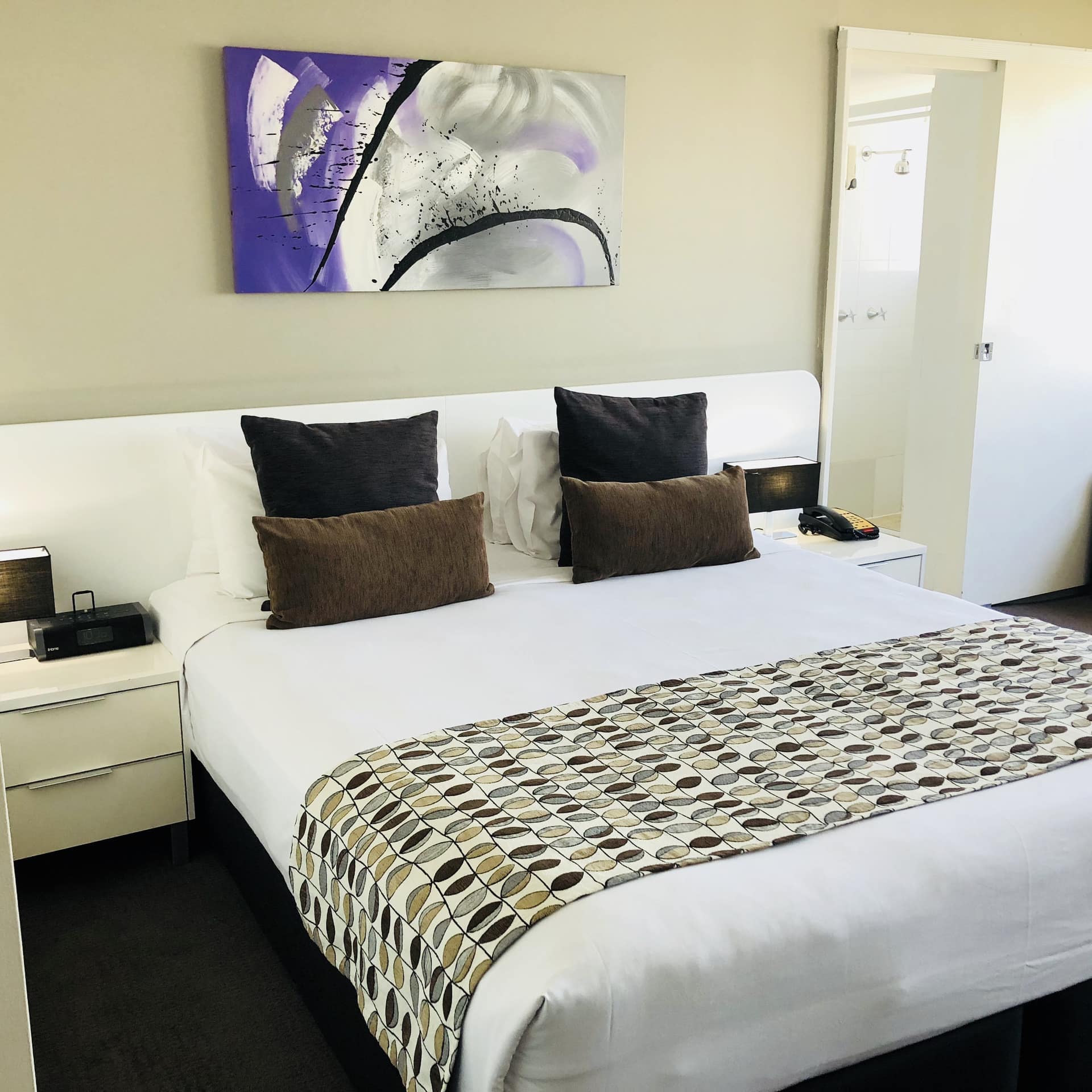A bedroom with black carpet, beige walls, a king size bed with white and brown bedding, and an abstract painting.