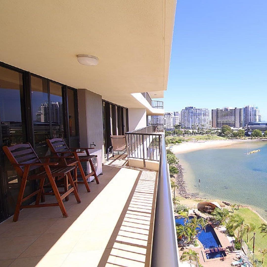 Covered balcony overlooking the ocean and beach with beige tiles and black railing and wooden lounge chairs.