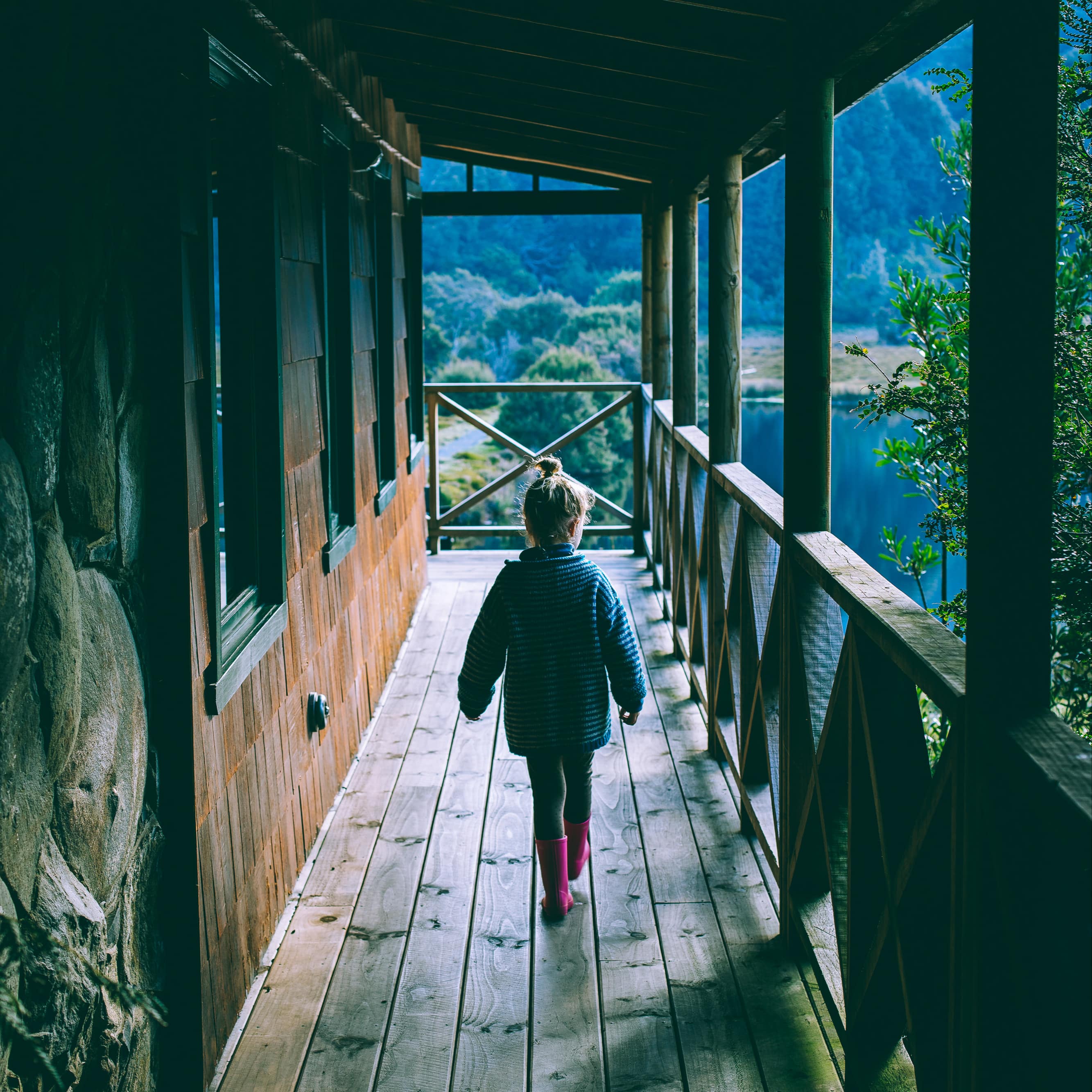 Young girl wearing a jacket and pink galoshes walks on wood wraparound porch of chalet with wood shake siding and lake view