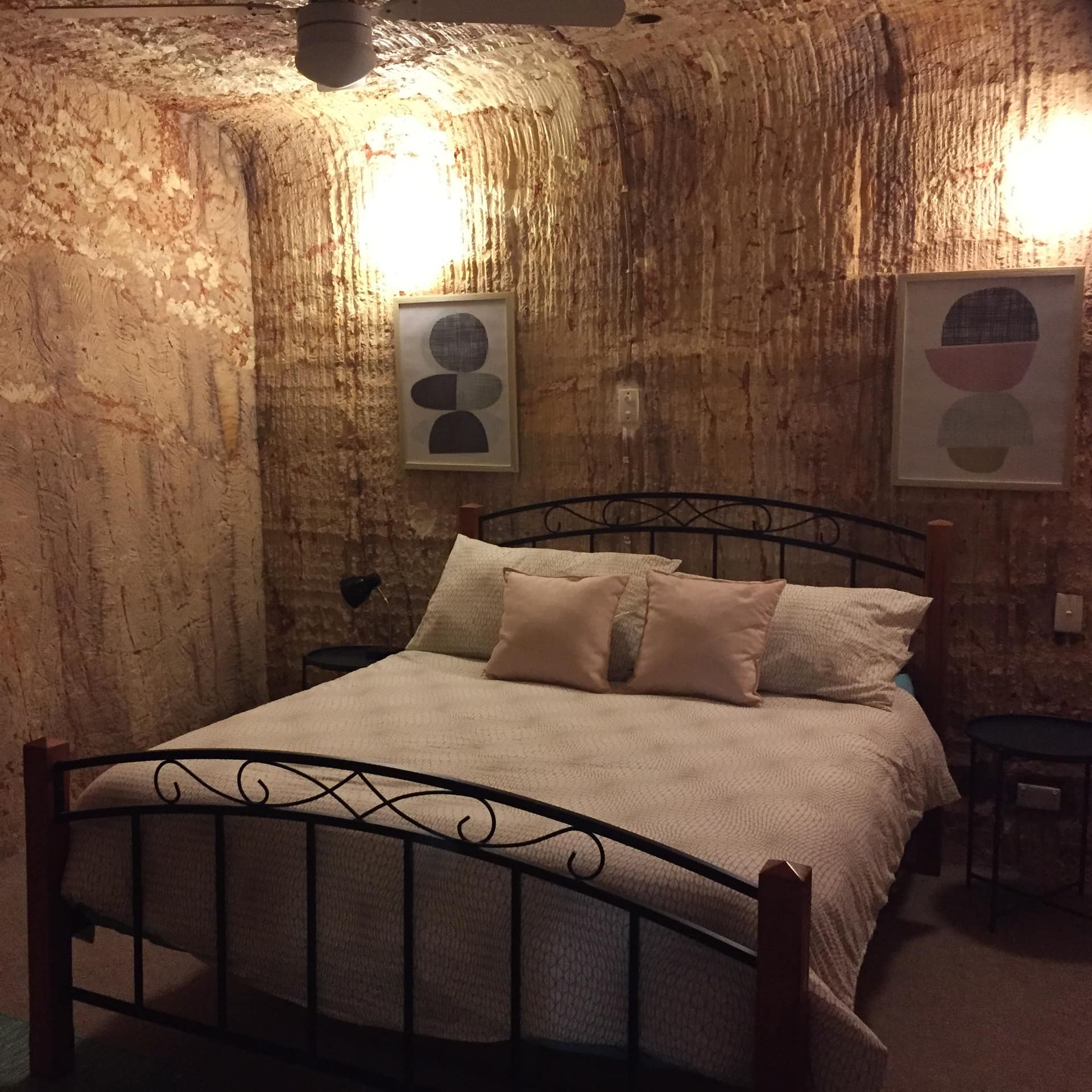 Underground bedroom with dugout walls, black metal king size bed with tan and light pink bedding, abstract art.