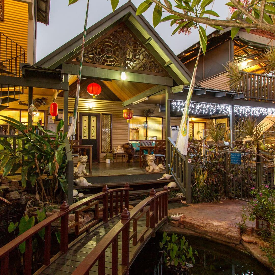 Exterior view of an East Asian-inspired home with a covered porch, a pond with a wooden bridge over it, thick vegetation.