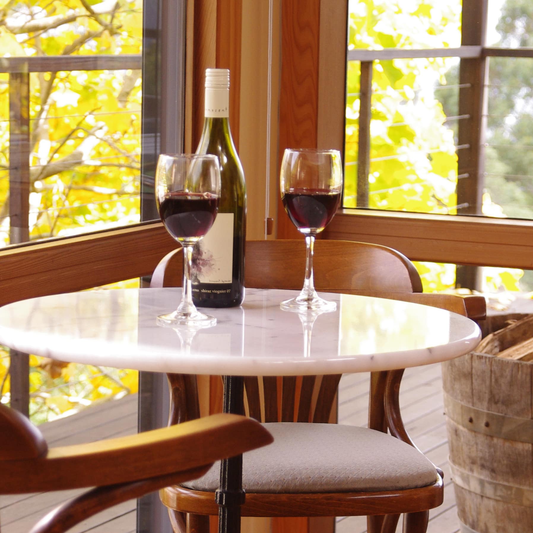 Wine glasses and red wine sit on a marble table with wood chairs in front of wide windows with nature views.