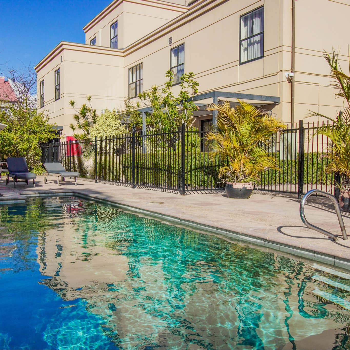 View of a pool, beige tile pool deck, outdoor dining set and lounge chairs, next to a large beige building.