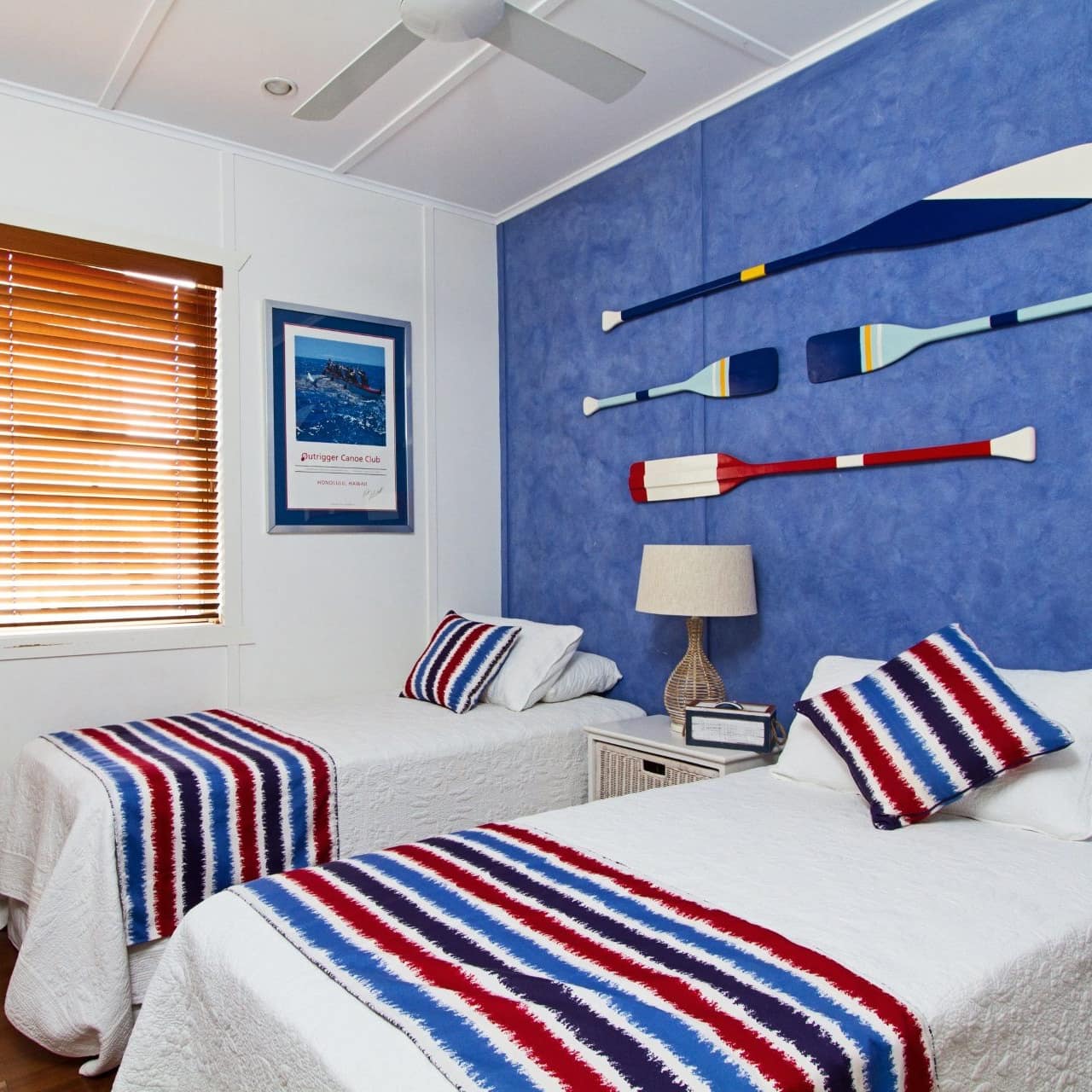Bedroom furnished in red, white, and blue with hardwood floors, blue accent wall, two twin beds, nautical decor.